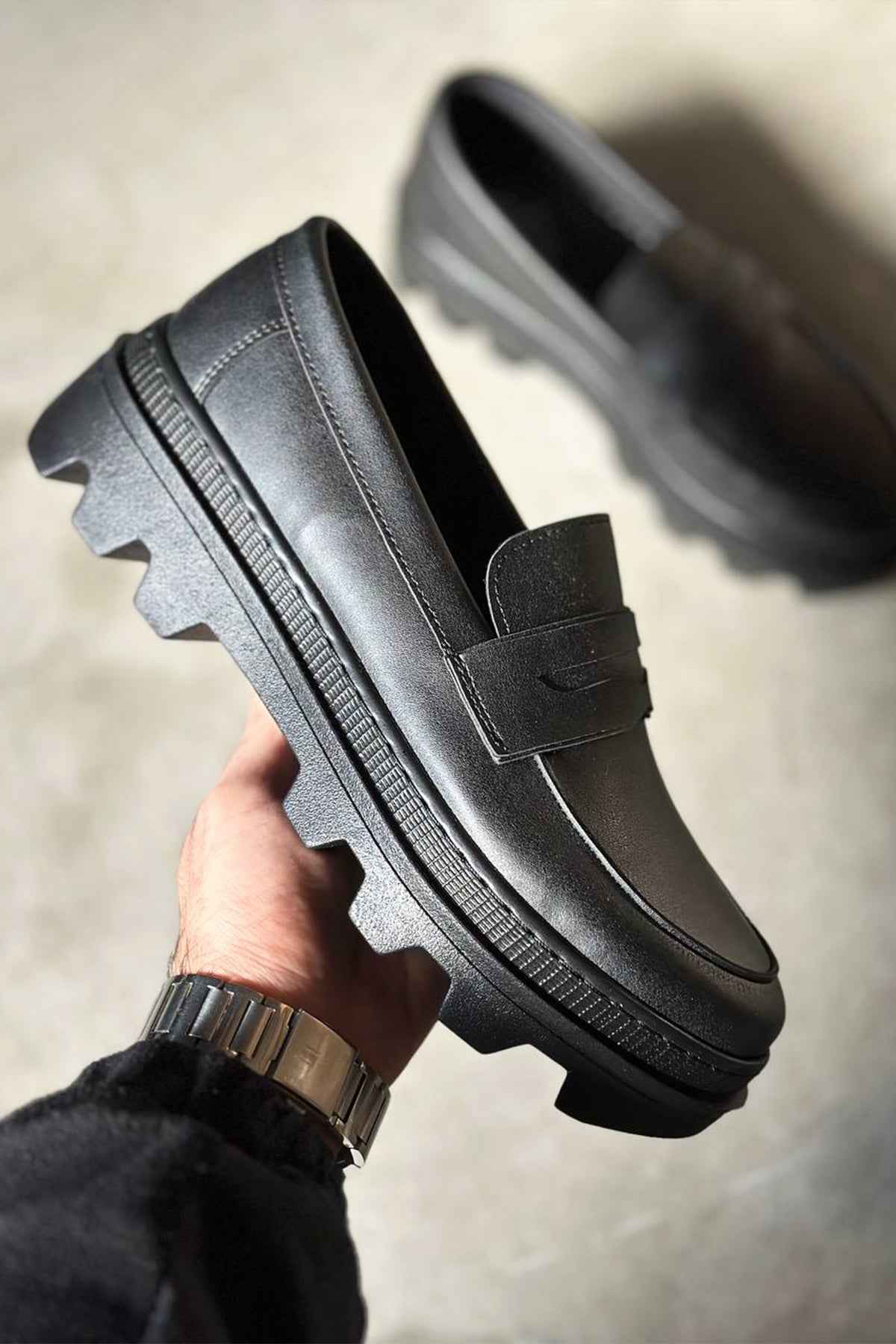 Men's black loafer in hand with strap, stitched seams, and tread sole