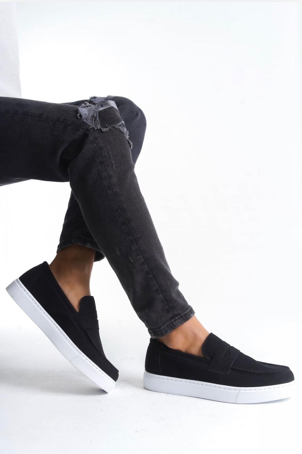 Men's black suede slip-on loafers with white sole, worn with dark jeans
