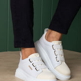White low-top sneakers with side buckles and stitched toe cap, worn with dark pants.