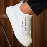 White low-top sneaker showing padded collar, side buckle straps, and logo text.