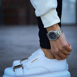 Men's white sneakers with buckle strap on leather upper, worn with dark pants.