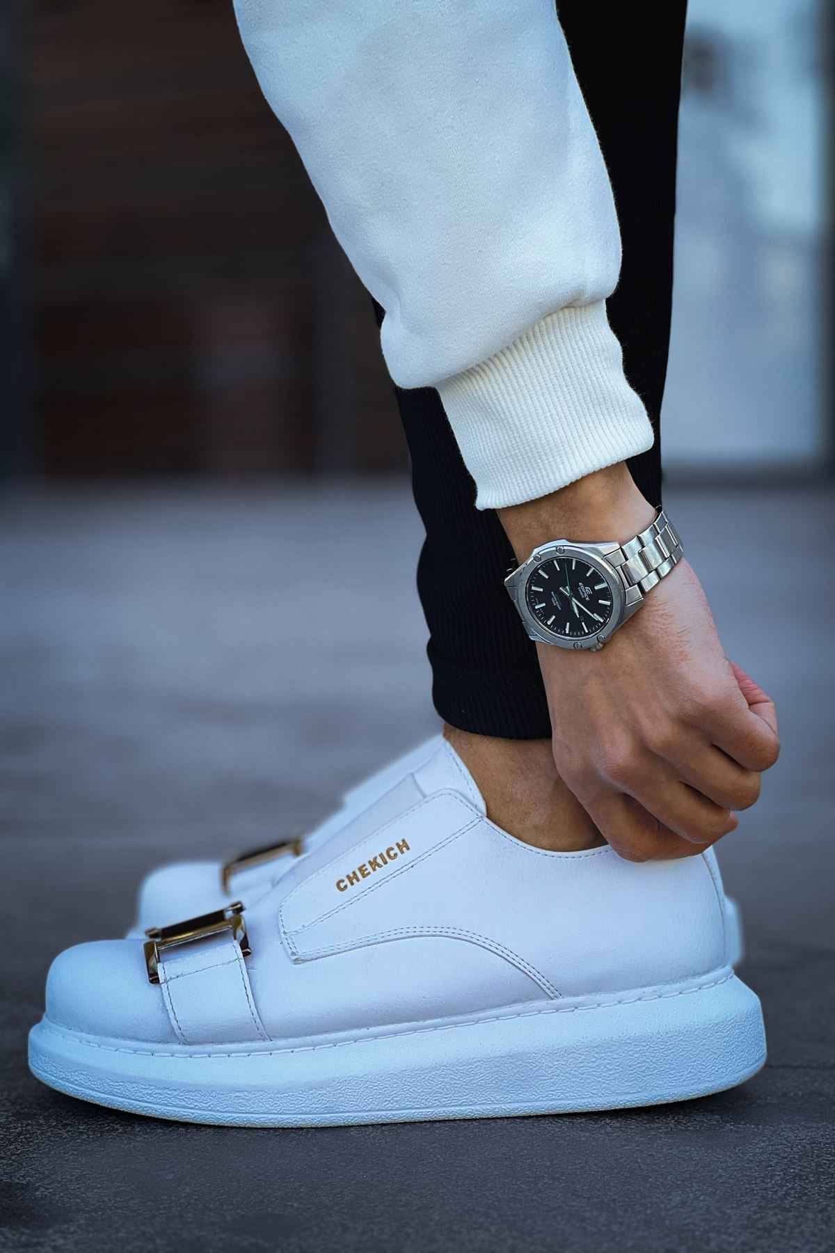 Men's white sneakers with buckle strap on leather upper, worn with dark pants.