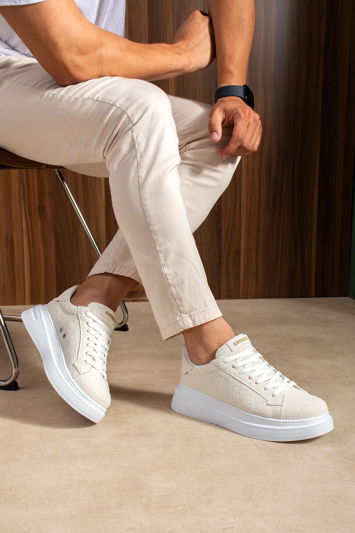 White suede lace-up sneakers on seated legs, with blue-gray rubber outsole.