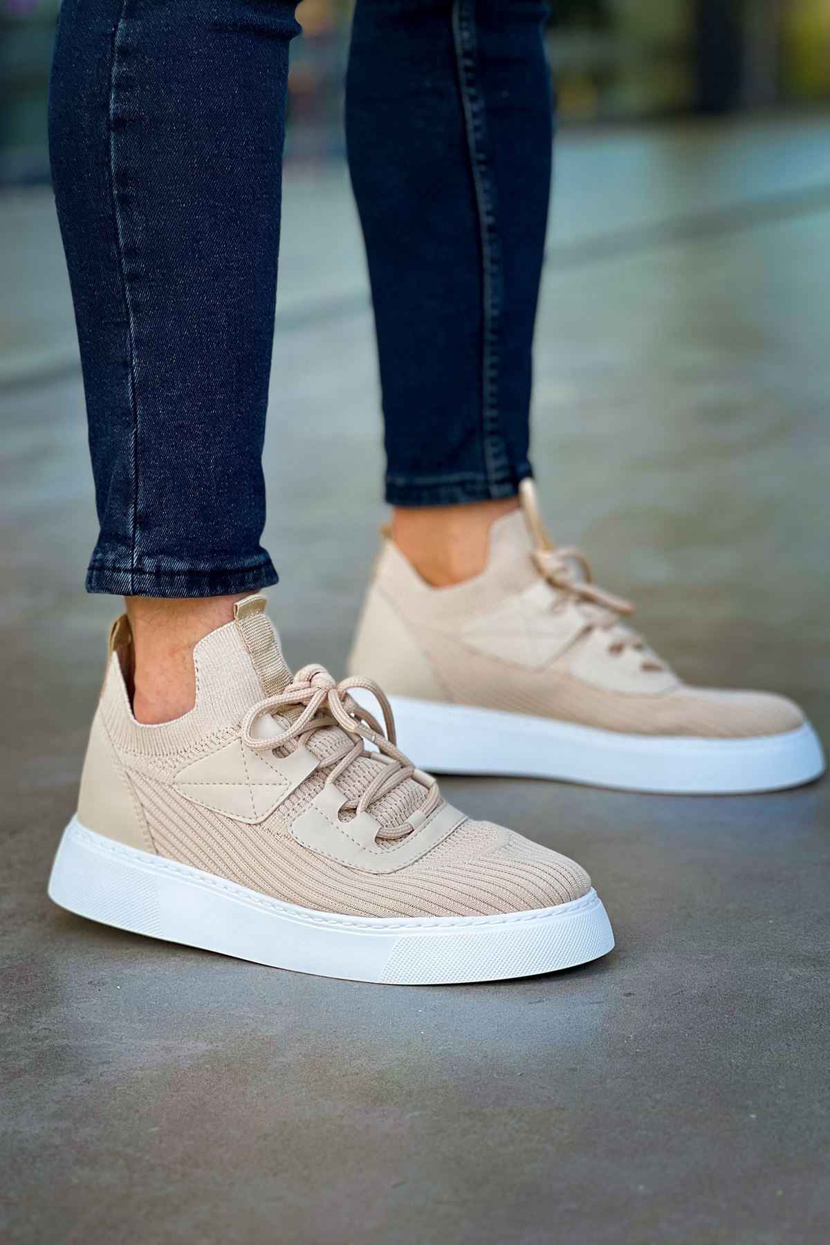 Beige knit sneakers with lace-up front and white rubber sole, worn with dark jeans.