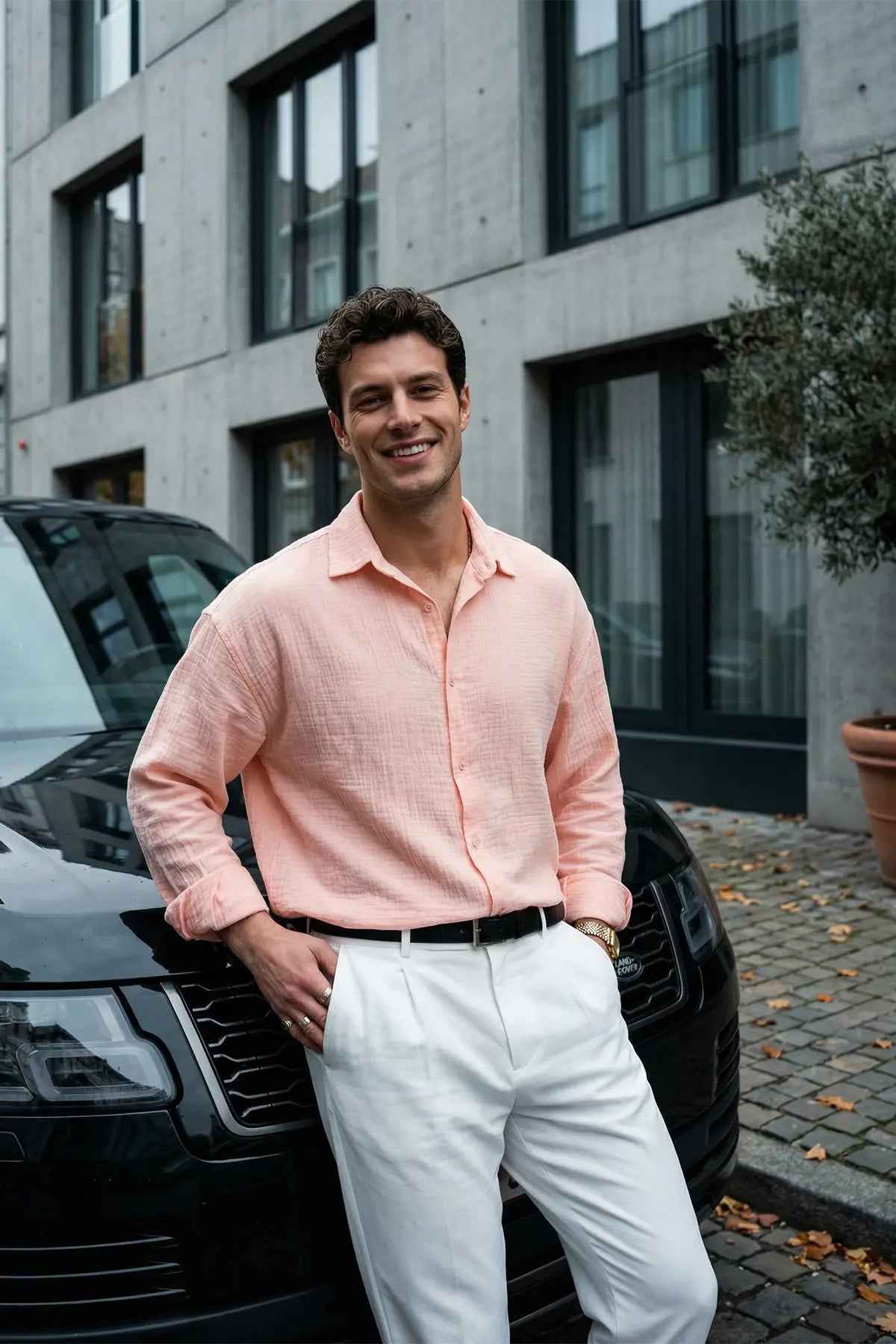 Mens pink dress shirt with button front, spread collar, long sleeves, worn untucked with white pants.