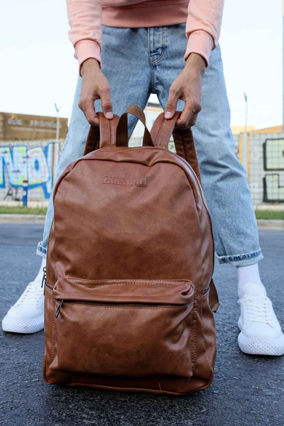 Tan leather backpack with front zipper pocket and top handle, held in hands