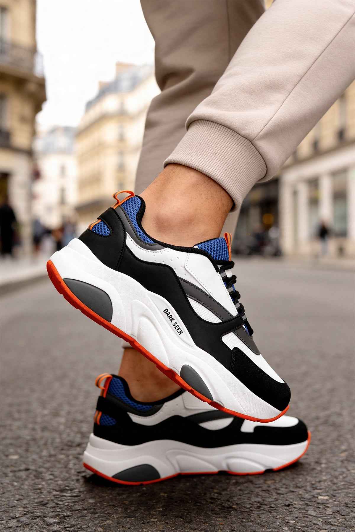 Men's lace-up sneakers on foot: white, black, and blue upper with orange-trim outsole.